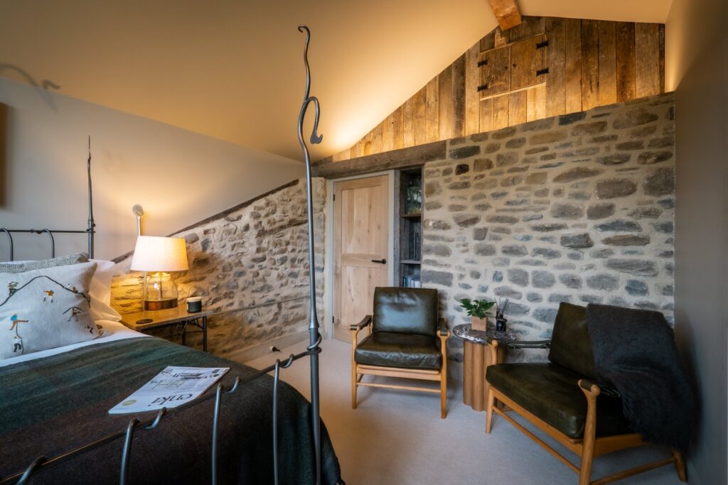 Bedroom with a double bed side on, a seating area with two seats and an exposed stone wall with led lighting