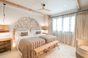 A twin bedroom with terracotta and beige bed linen, a recessed arch with a feature stone wall, voile and curtain dressings for the windows