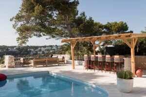 The outdoor terrace of a Menorcan Villa with a large pergola housing a fully furnished outdoor kitchen and bar area and a separate seating area for 8 to 10 people