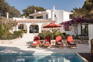 A view from the outdoor terrace looking back across the pool and sun loungers at the Menorcan villa with a terracotta tile roof and beautiful arched colonnade