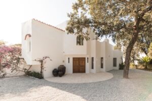 A wide angle view of a crisp white Menorcan villa, simple gravel driveway, with large pots outside the handmade entrance doors