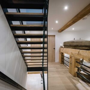 Hallway view of 1 flight of stairs with a juxtaposition of materials using a metal frame, wood treads and glass panelling