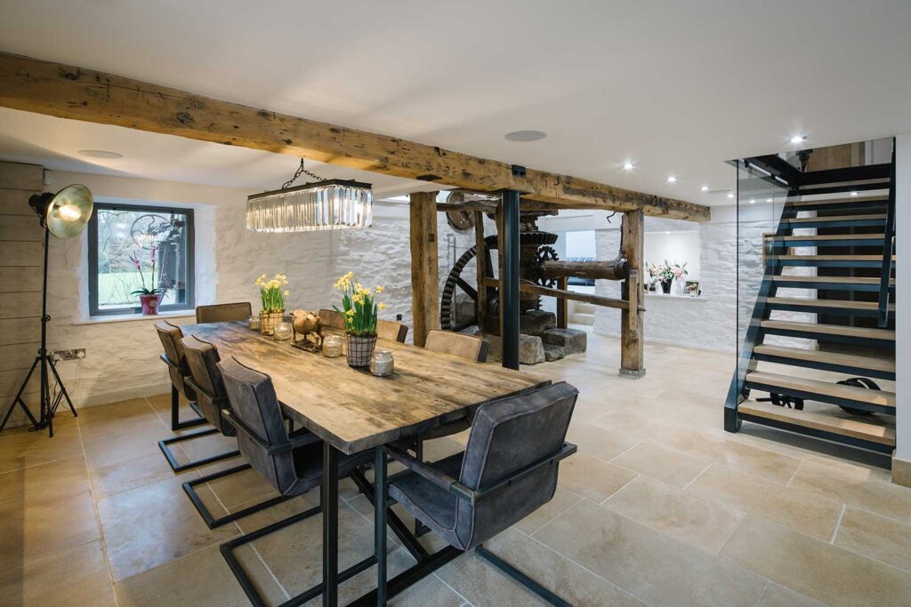 Dining room of a Grade II Mill restoration with natural stone flooring and reclaimed timber dining table with a bespoke glass, oak and metal staircase