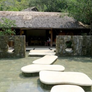 Salutogenic design vista of large irregular shaped stepping stones over a pond leading to a wooden structure with a thatched roof