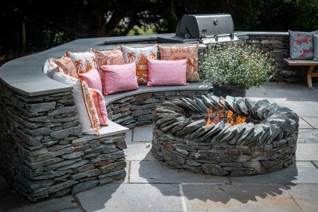 Bespoke craftsman stone fire pit and stone curved seating bench with vibrant pink and orange cushions on the rear patio of a beautiful home renovation designed by Charlotte Findlater Design, an award winning interior design studio specialising in luxury, high-end residential & commercial projects that respect time, nature and people.
