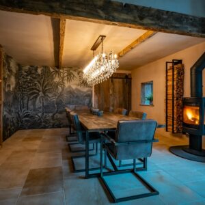 A whole dining room view showing a lights on chandelier, a large reclaimed wood dining table with 10 chairs and a lit log burner