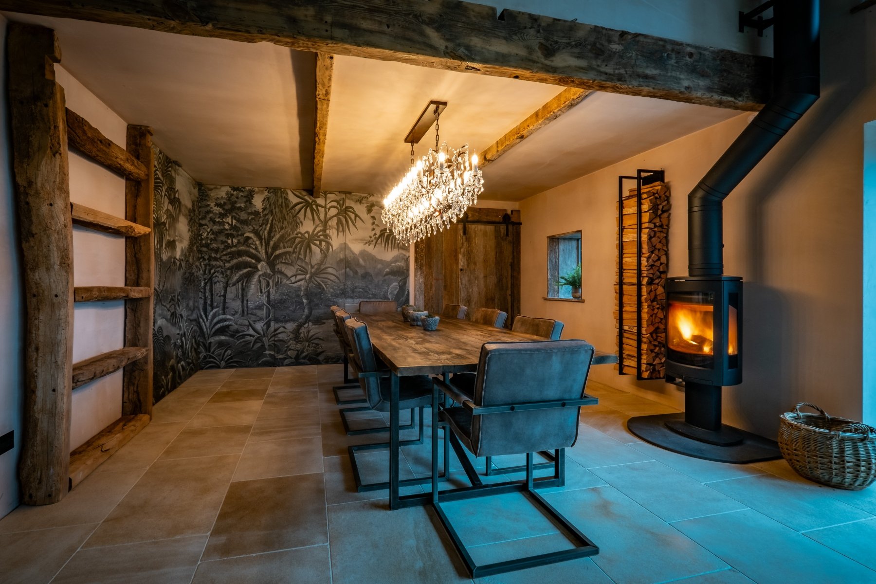 A whole dining room view showing a lights on chandelier, a large reclaimed wood dining table with 10 chairs and a lit log burner