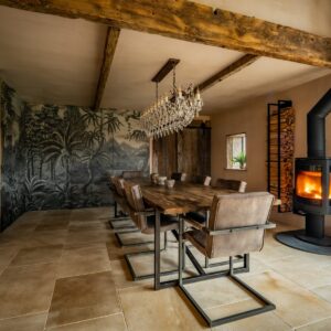 A dining room view drenched with natural light, focusing on a large reclaimed wood dining table, a jungle screen wall feature decoration and a lit log burner