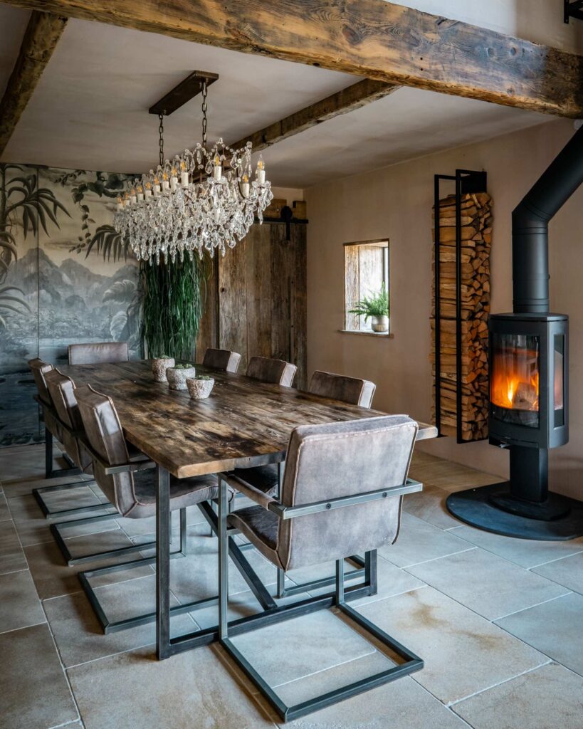 A dining room view drenched with natural light, focusing on a large reclaimed wood dining table, a jungle screen wall feature decoration and a lit log burner