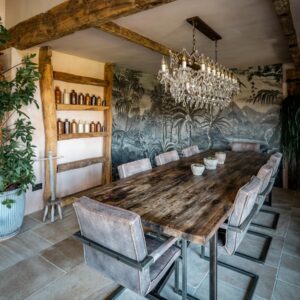A biophilic designed dining room view drenched with natural light, focusing on a large reclaimed wood dining table, a rock crystal chandelier, jungle screen wall feature decoration and a bespoke beam shelf with vintage ink bottles