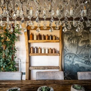 A biophilic designed dining room portrait view of a rock crystal chandelier, a large reclaimed wood dining table with three stone pots with living moss and a bespoke beam shelf with vintage ink bottles