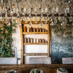 A biophilic designed dining room portrait view of a rock crystal chandelier, a large reclaimed wood dining table with three stone pots with living moss and a bespoke beam shelf with vintage ink bottles